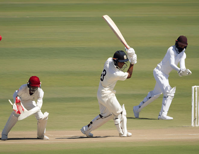 Batsman swings the cricket bat forcefully, connecting with the ball, which soars high into the clear blue sky, while fielders chase after it.
